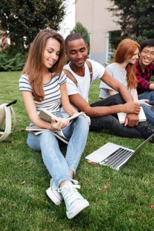 Multiethnic group of happy young people reading book and using laptop on lawn outdoors